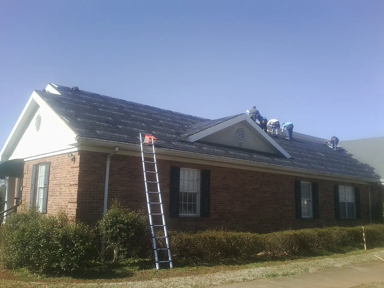 Crew tearing shingles off a brick ranch roof with a ladder set — Metal Roof Repair work by our crew in Mathews